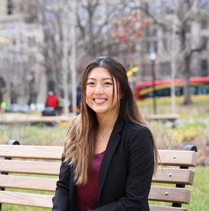 MSU alumna Angela Yuan smiles while sitting on a wooden bench
