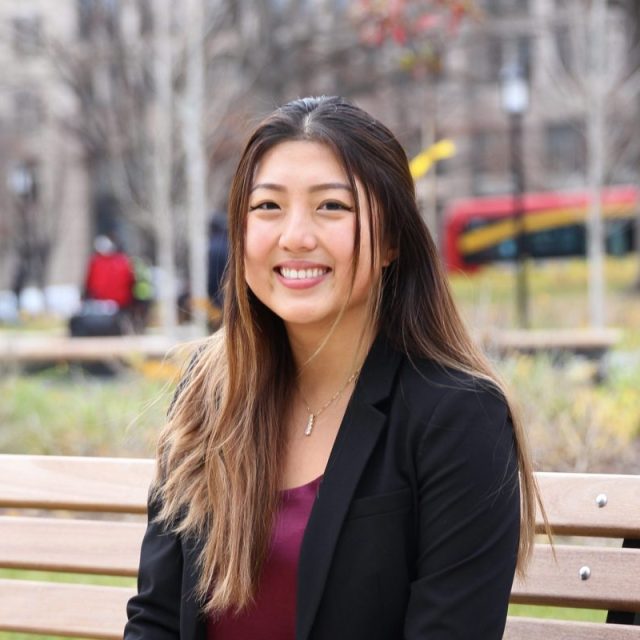 Image of Angela Yuan in a black blazer and dark red top, sitting outside on a wooden bench and smiling at the camera.
