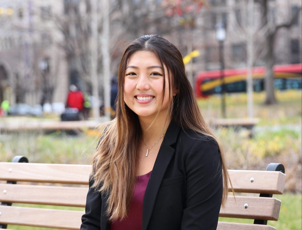 Image of Angela Yuan in a black blazer and dark red top, sitting outside on a wooden bench and smiling at the camera.