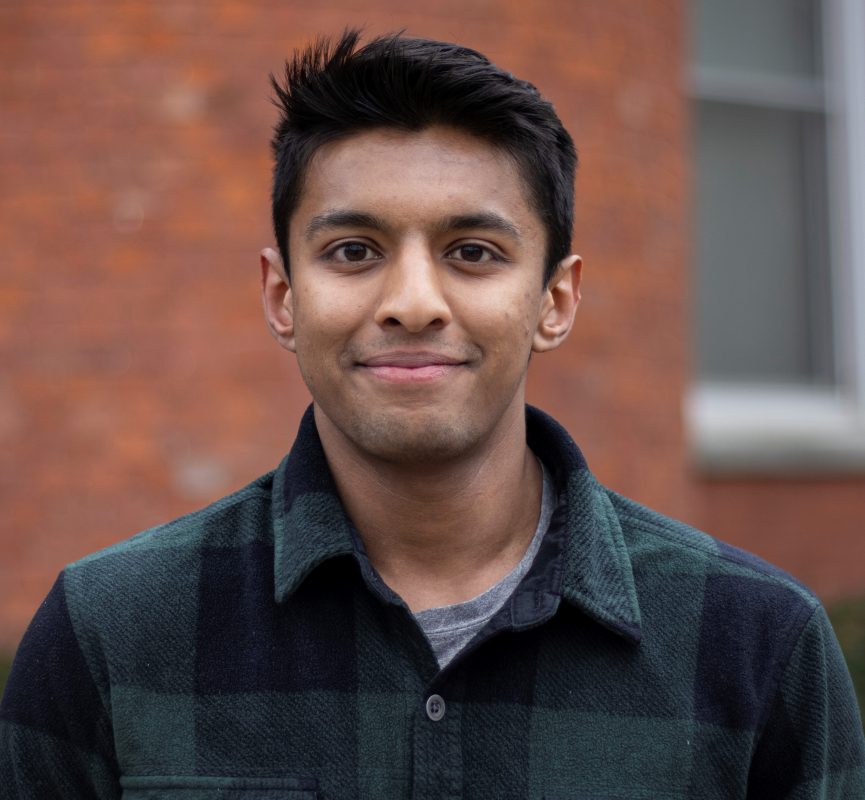 MSU Honors College alumnus Aaron Philip wearing a plaid green shirt and standing in front of a red brick building.
