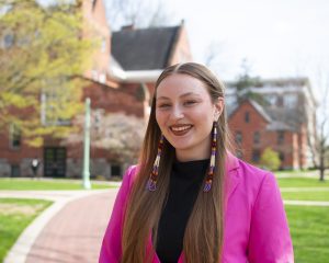 Headshot of a student in a hot pink blazer with long beaded earrings, with a red brick path and two red brick buildings in the background.