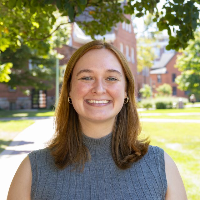 Headshot of Caroline Downes in a polished ribbed tank, with greenery and red brick buildings in the background.