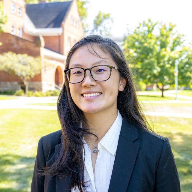 Headshot of Grace Urban in a white collared shirt and black blazer, with greenery and a red brick building in the background.