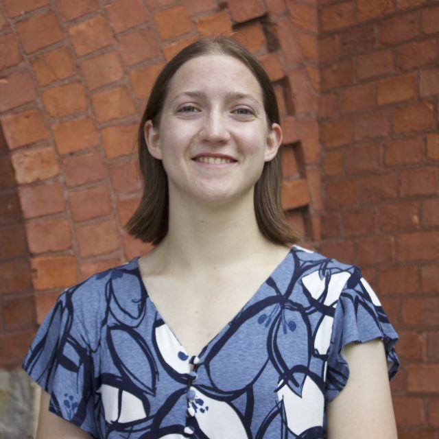 Headshot of Rachel Schenck in light purple top with white and black oval designs. The background is a red brick wall.