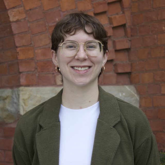 Headshot of Sydney Logsdon in a white shirt and olive green blazer. The background is a red brick wall with a row of tan brick.