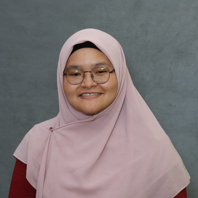 Professional headshot of Nur Aminah Benti Halim, who is wearing a light pink head covering and dark red top. Nur is smiling against a gray backdrop.