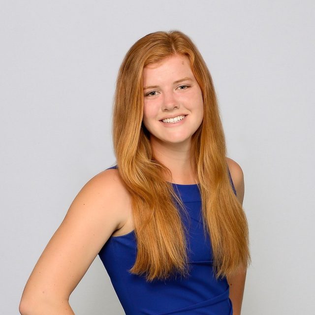 A professional photo of Caroline Colby Orcutt from the waist up, against a plain grey background. Orcutt is smiling and wearing a vibrant blue top.