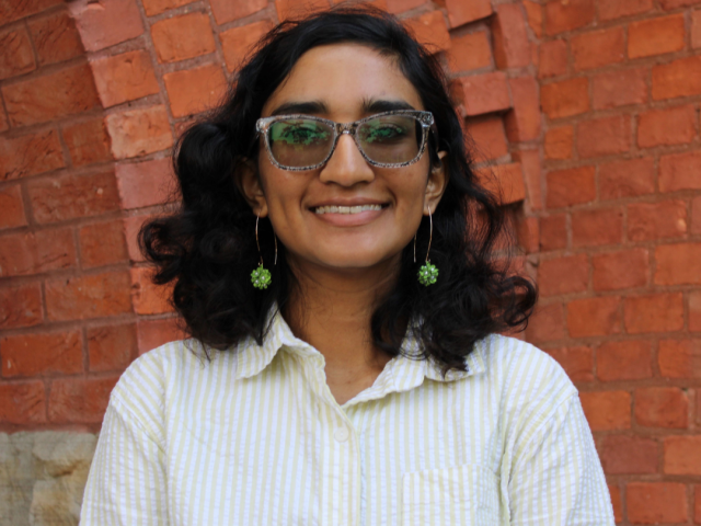 Headshot of Poulomi Dey standing against a red brick background, wearing a yellow and white striped collared shirt and gold hoop earrings with clusters of green flowers on the bottom.