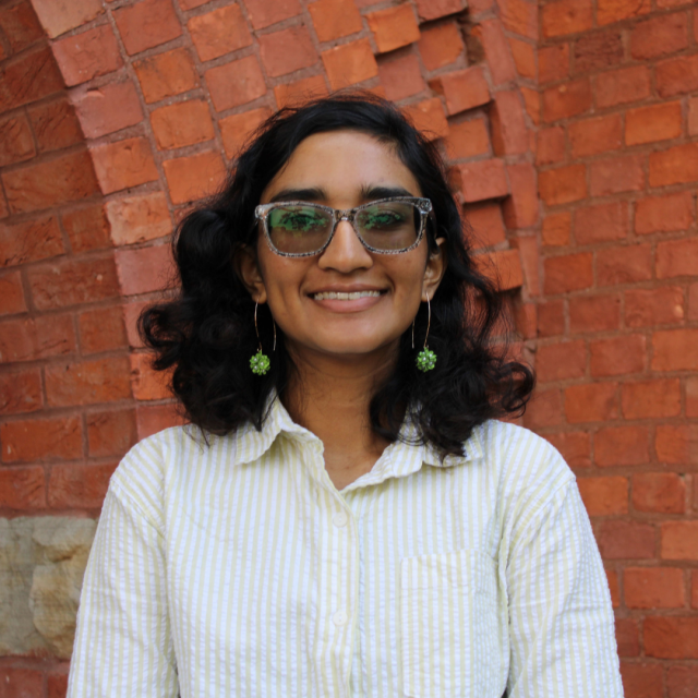 Headshot of Poulomi Dey standing against a red brick background, wearing a yellow and white striped collared shirt and gold hoop earrings with clusters of green flowers on the bottom.