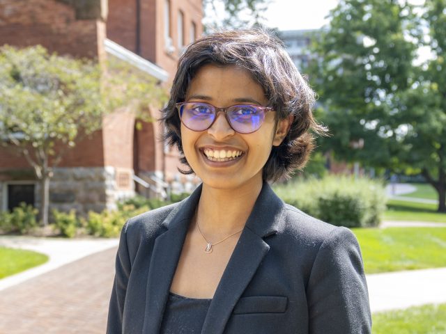 Professional headshot of Veona Cuthino in a black top and black blazer. In the background is a red brick building, a brick path, and lush greenery.