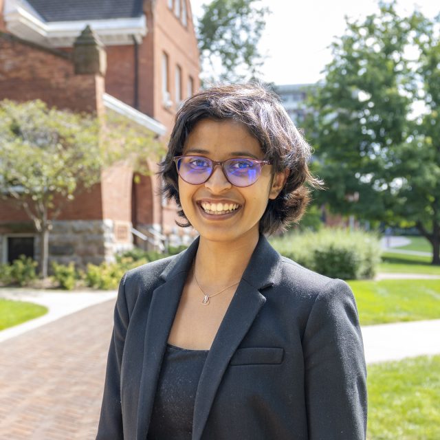 Professional headshot of Veona Cuthino in a black top and black blazer. In the background is a red brick building, a brick path, and lush greenery.