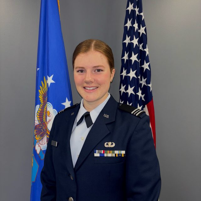 Caroline (Colby) Orcutt in a formal U.S. Air Force uniform stands in front of two flags, one on each side. The U.S. flag with stars and stripes is on the right, and the Air Force flag with an eagle emblem is on the left. Orcutt has a slight smile and looks directly at the camera. The uniform features various ribbons and insignia on the chest. The background is a plain gray wall.