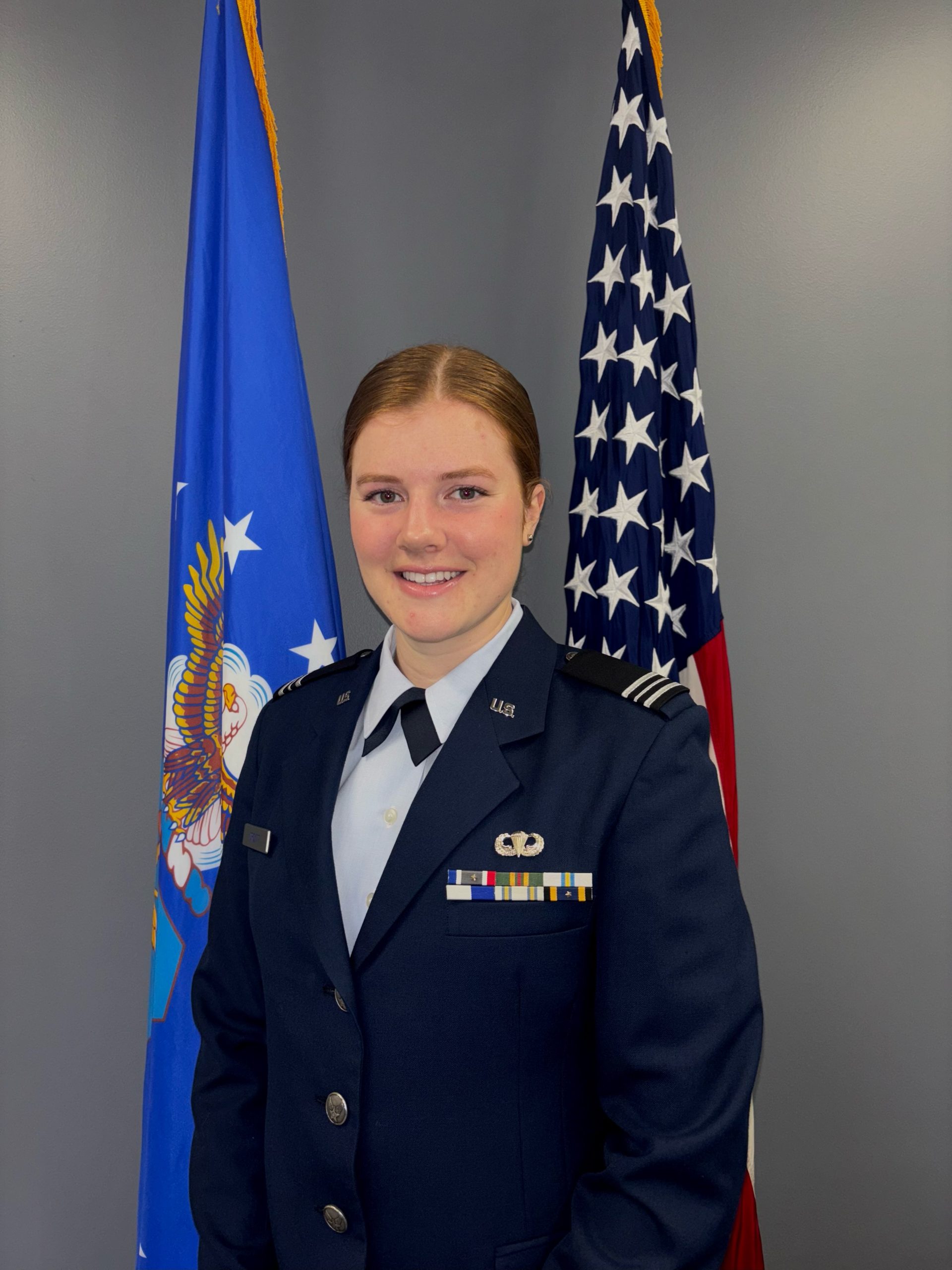 Caroline (Colby) Orcutt in a formal U.S. Air Force uniform stands in front of two flags, one on each side. The U.S. flag with stars and stripes is on the right, and the Air Force flag with an eagle emblem is on the left. Orcutt has a slight smile and looks directly at the camera. The uniform features various ribbons and insignia on the chest. The background is a plain gray wall.