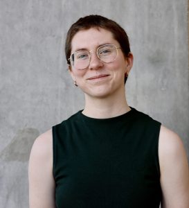 Sydney Logsdon is smiling warmly at the camera. Logsdon is wearing a sleeveless dark green top and gold hoop earrings. The background is a plain gray concrete wall.