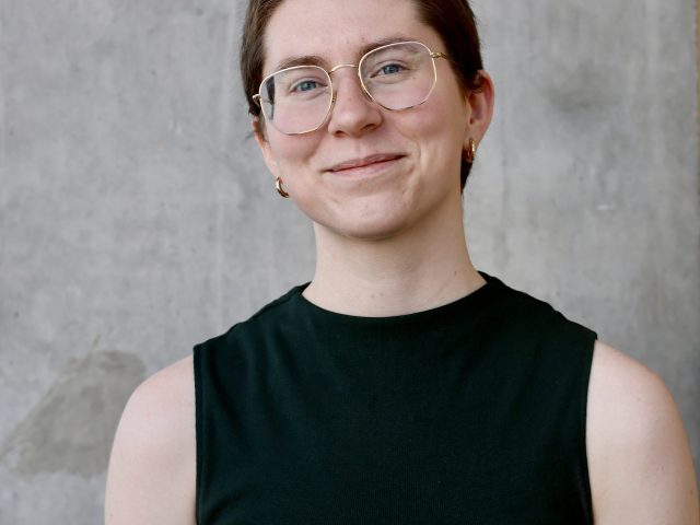 Sydney Logsdon is smiling warmly at the camera. Logsdon is wearing a sleeveless dark green top and gold hoop earrings. The background is a plain gray concrete wall.