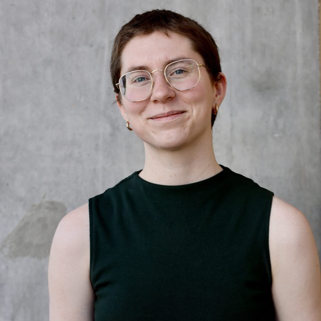 Sydney Logsdon is smiling warmly at the camera. Logsdon is wearing a sleeveless dark green top and gold hoop earrings. The background is a plain gray concrete wall.