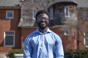 Jerome Hamilton Jr. is smiling at the camera. Jerome is wearing a blue button down shirt and standing in front of a red brick building.