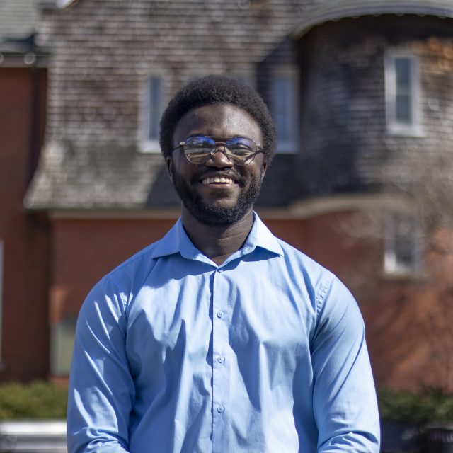 Jerome Hamilton is smiling at the camera. Jerome is wearing a blue button down shirt and standing in front of a red brick building.