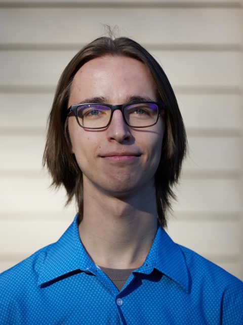 Erik Brodsky is smiling at the camera, wearing a blue button down and standing in front of a white wall.