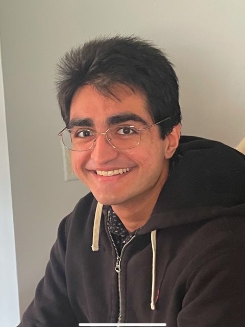 Jesse Sanhu is smiling at the camera and is wearing wire framed glasses and a black zip-up.