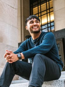 Image of Mustafa Syed sitting on steps in front of a stone building. Syed is wearing a dark knit sweater and dark pants, smiling and looking off to the side.