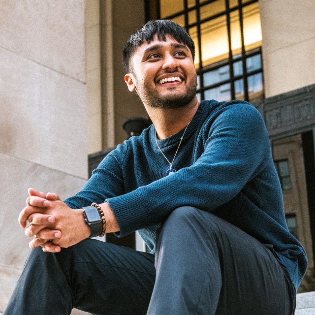 Image of Mustafa Syed sitting on steps in front of a stone building. Syed is wearing a dark knit sweater and dark pants, smiling and looking off to the side.