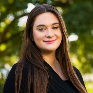 Claire Smith is standing in front of a nature background, wearing a black blazer, and smiling at the camera.