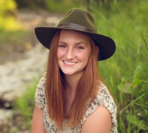 Isabelle Radakovich in front of a nature background, wearing a green hat and floral top, smiling at the camera.