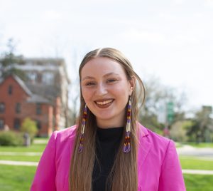 Kaylin Casper is standing in front of a red brick building, wearing a pink blazer, and smiling at the camera