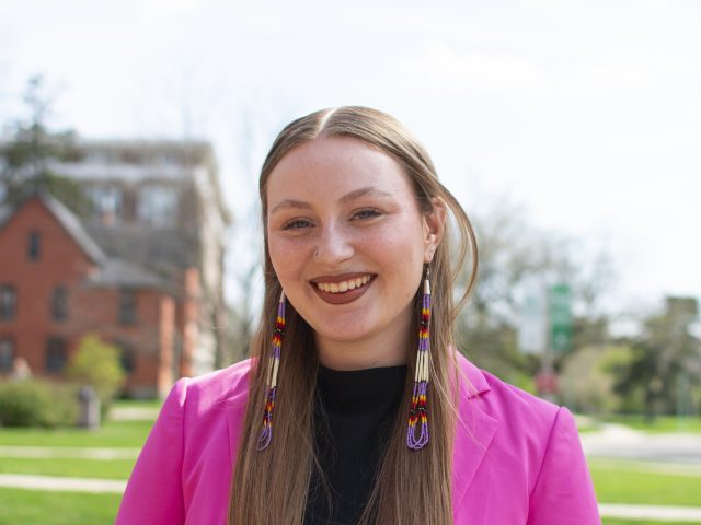 Kaylin Casper is standing in front of a red brick building, wearing a pink blazer, and smiling at the camera