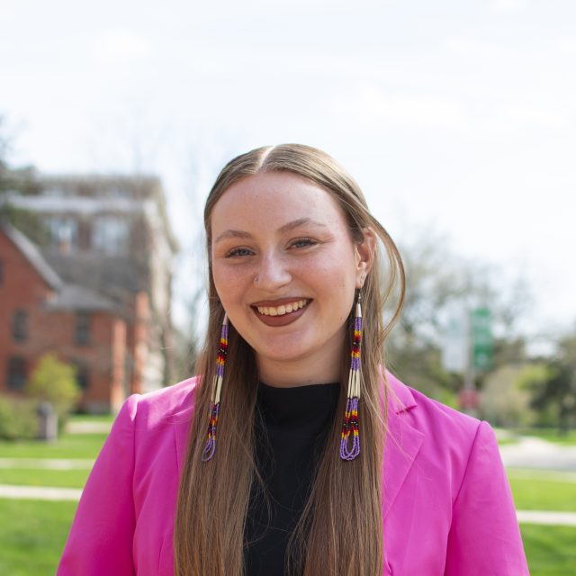 Kaylin Casper is standing in front of a red brick building, wearing a pink blazer, and smiling at the camera