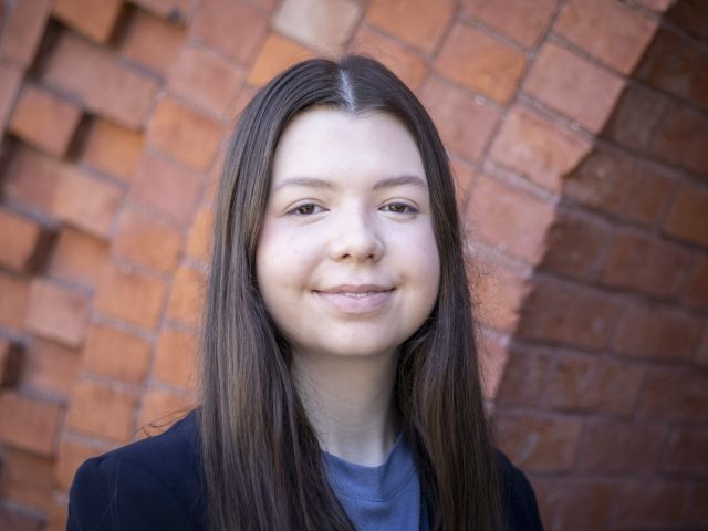 Eleanor Pugh smiles at the camera, wearing a black blazer and standing in front of a brick wall.