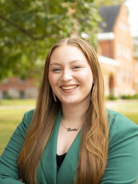 Image of Michigan State University student Kaylin Casper smiling in a green blazer and black top. There is a red brick building and greenery in the background.