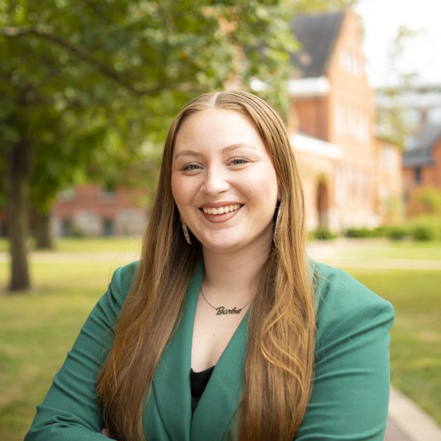 Image of Michigan State University student Kaylin Casper smiling in a green blazer and black top. There is a red brick building and greenery in the background.
