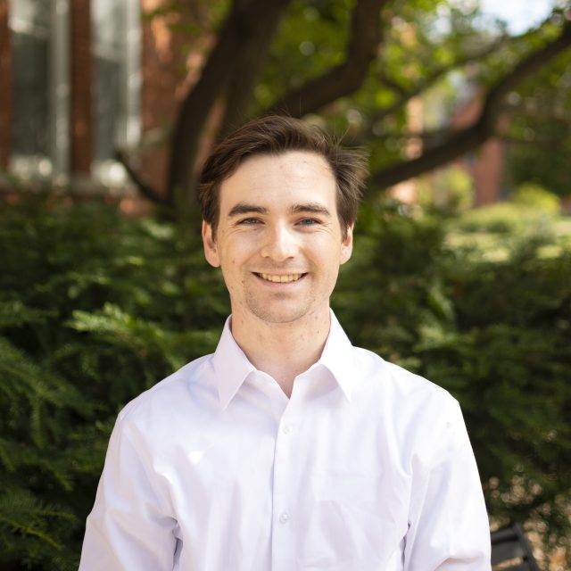 Image of Michigan State University student William Castanier smiling in a white collared shirt, with greenery and a red brick building in the background.