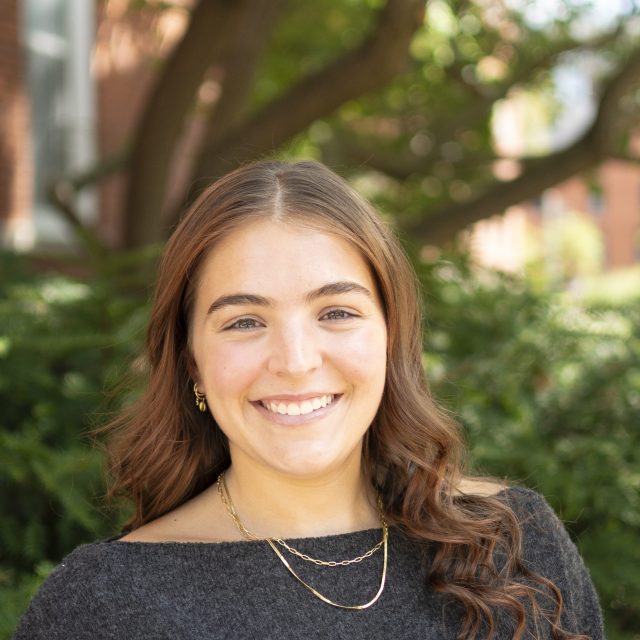 Image of Michigan State University student Julia Preservati smiling, while wearing a dark top and thin gold necklaces. There is greenery and a red brick building in the background.