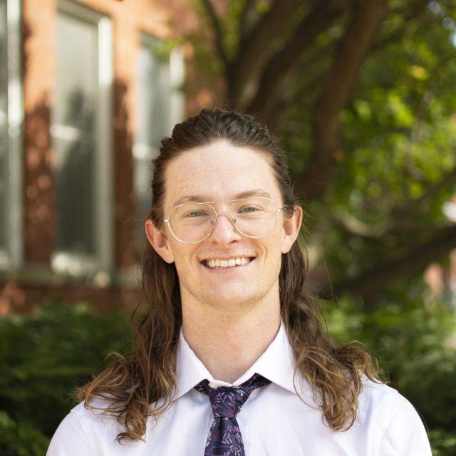 Image of Michigan State University student Colin Reese smiling in a white collared shirt and dark necktie. There is a red brick building and greenery in the background.