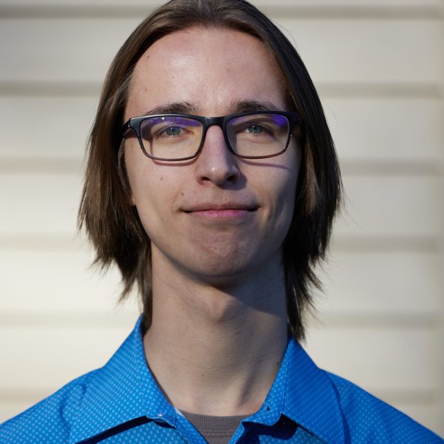 Erik Brodsky is wearing a blue collared shirt and glasses and is smiling at the camera.