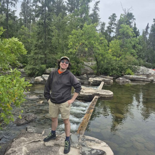 Image of Karter Burgdorf smiling at the camera standing on a rock on a river. Burgdorf is wearing a dark hoodie and khaki shorts.