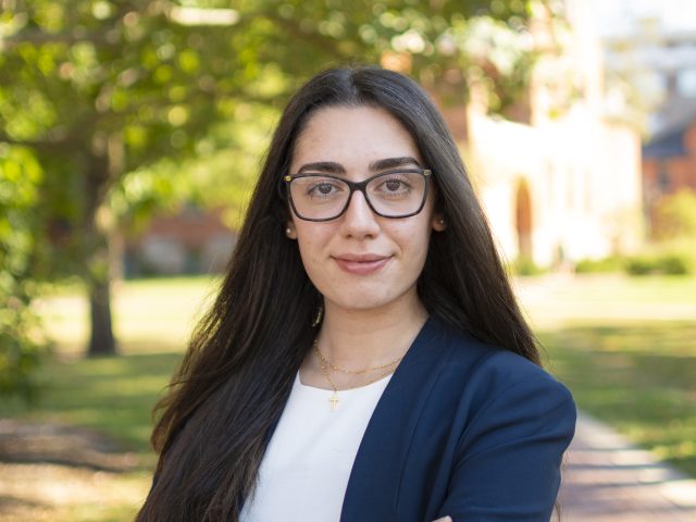 Sarah Krikor is standing in front of a nature background and is wearing a blazer and glasses.