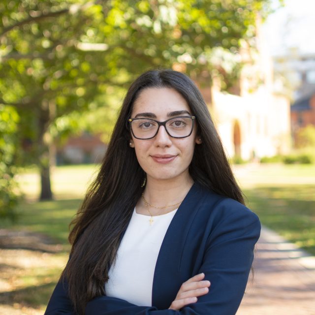 Sarah Krikor is standing in front of a nature background and is wearing a blazer and glasses.