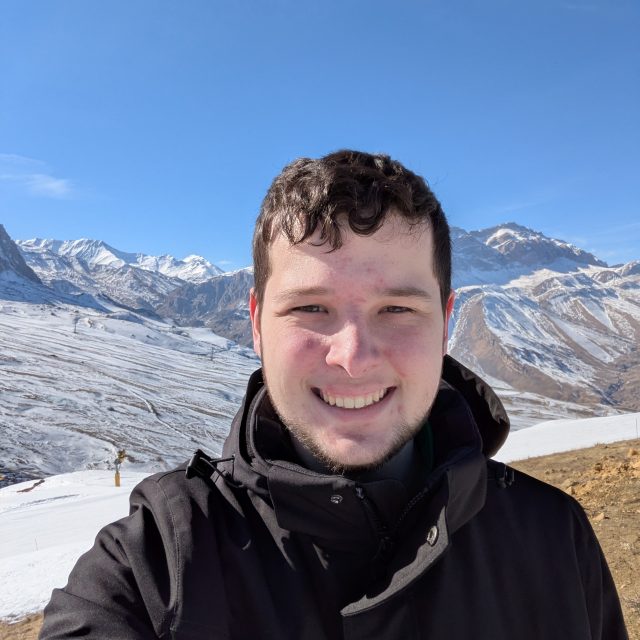 Image of Adam Peck smiling at the camera and standing in front of a snowy mountain range, wearing a black jacket.