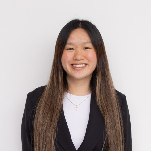 Image of Tammy Thai smiling and standing in front of a white wall, wearing a white shirt and black blazer.