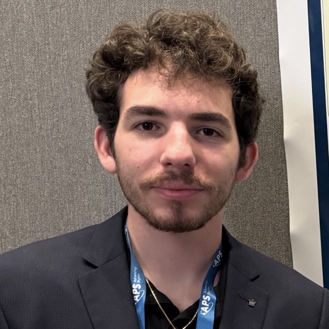 Image of Michigan State University student Kevin Eisenberg softly smiling and standing against a wall in a black suit jacket, black polo, gold necklace, and blue lanyard.