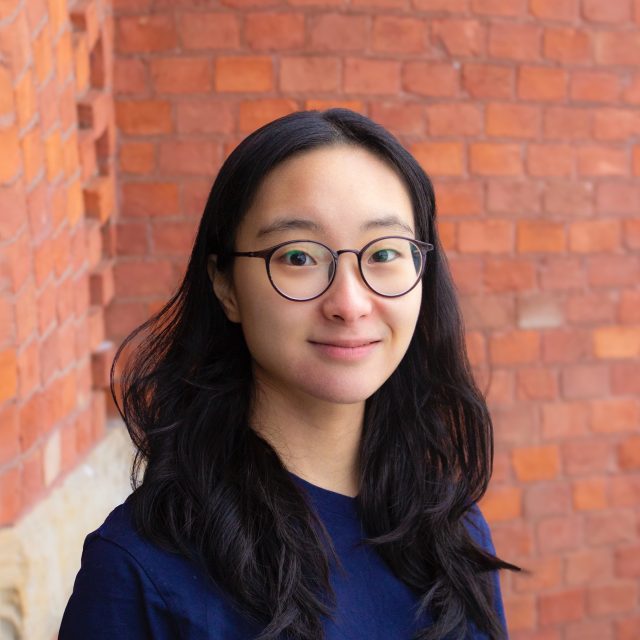 Image of Michigan State University student Lauren Jin smiling softly in a navy shirt and brown glasses. A red brick wall is in the background.