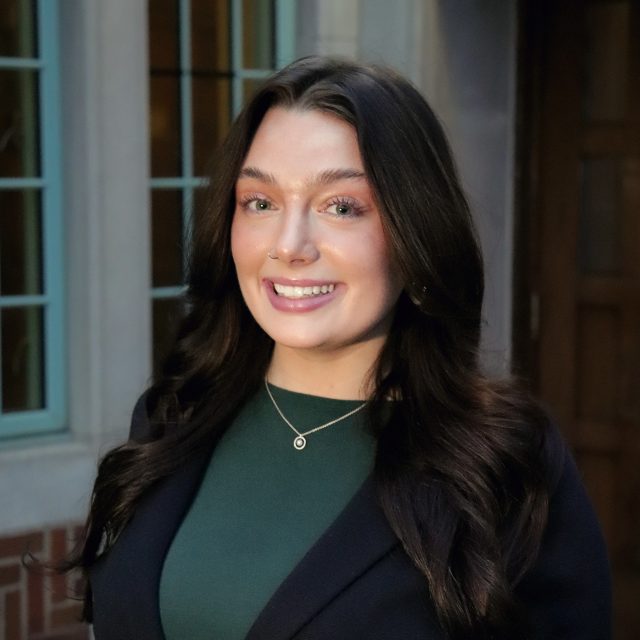 Professional photo of Michigan State University Honors College student Lily Dixon smiling in a dark green top, black blazer, and small pendant necklace. A dark brick building with teal-trim windows is in the background.