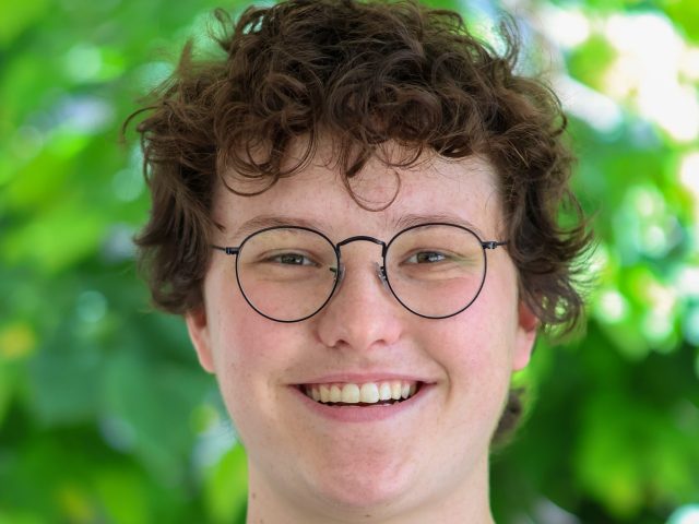 Emma Andrechek is smiling at the camera, wearing a black shirt and wire framed glasses, with a nature background.