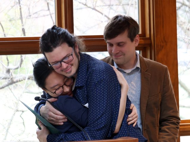 Image of Michigan State University Honors College student Lauren Jin and faculty member Pablo Giuliani in a warm embrace at a mentor appreciation event in Cowles House. Giuliani is holding a green award certificate, and faculty member Kyle Godbey is in the background.