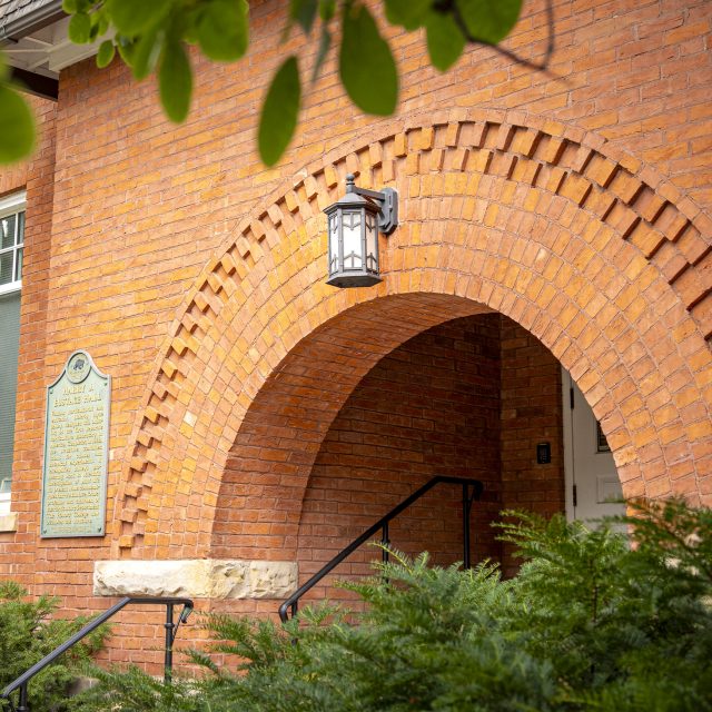 Image of Eustace-Cole Hall on Michigan State University's campus. The red brick building has an arched entrance and is surrounded by lush greenery.