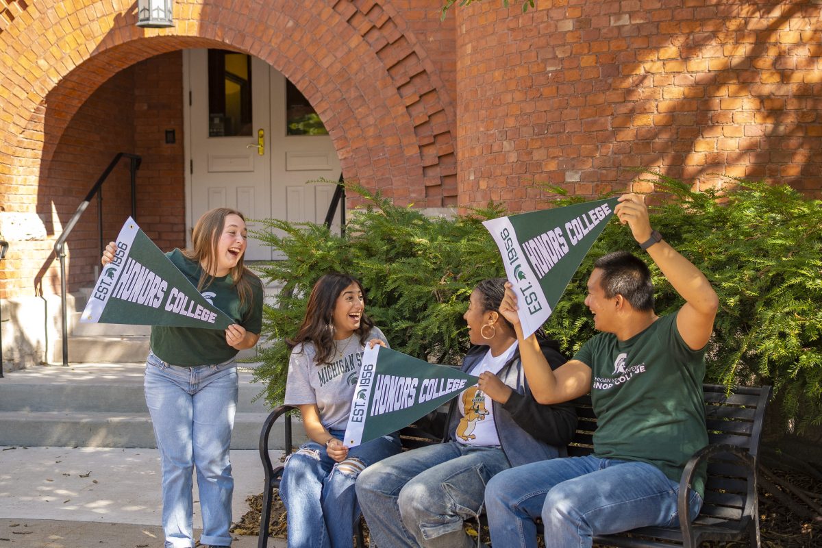 A group of four MSU Honors College students in front of the red brick entrance to Eustace-Cole Hall. Three students are sitting on a black bench, while another is standing. The students are in jeans and various MSU t-shirts, holding green and white Honors College pennants.